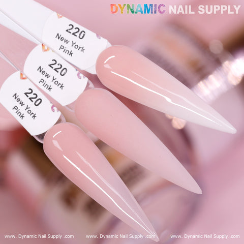 A close-up of three elongated nail samples showcases the New York Pink - Nude Acrylic Powder (#220) from Dynamic Nail Supply, known as the best cover color for daily usage. The nails display a soft, pale pink gradient that offers long-lasting wear. In the background, blurred nail polish bottles enhance the focus on these striking foreground samples.