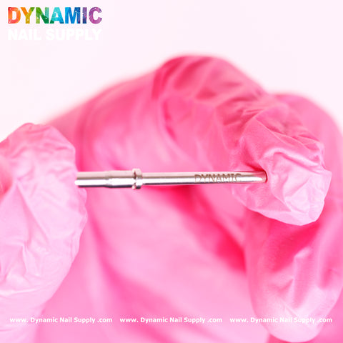 A hand in a pink glove holds a small, metallic nail drill bit, perfect for pairing with the Small 3.5mm Easy-Off Mandrel for Smaller Sanding Band. The text DYNAMIC and NATANIUM are engraved on the bit. Against a white background, Dynamic Nail Supply is printed in rainbow colors at the top left corner, with a website URL at the bottom.