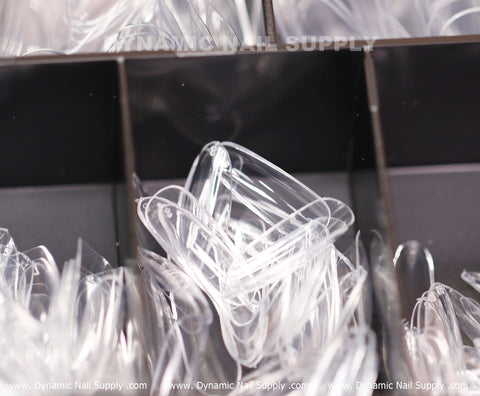 A close-up view showcases several transparent Round M Full Cover Tips - Soft Gel X tips neatly arranged in open compartments. These clear, slightly curved nail tips come in varying medium-long lengths, stacked and overlapping. In the background, the blurred logo of Dynamic Nail Supply is visible on the surface.