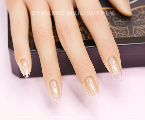 A mannequin hand showcases clear, glossy, medium-long press-on nails from the [Round M] Full Cover Tips collection by Dynamic Nail Supply, elegantly resting on a decorative box. These transparent soft gel X tips exude a modern and trendy style. The background features a soft pink surface with Dynamic Nail Supply subtly visible above the hand.