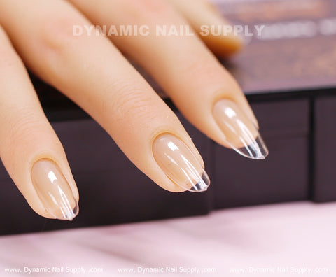 A close-up showcases a mannequin hand adorned with almond-shaped nails. These press-on tips feature a glossy, transparent tip that smoothly transitions into a natural nude hue. The backdrop is softly blurred, with the text "Dynamic Nail Supply" subtly visible above the fingers.