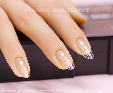 A close-up image features a mannequin hand displaying glossy, almond-shaped nails with clear tips and a beige base, created using the [Round M] Full Cover Tips - Soft Gel X tips. In the background, the blurred logo reads Dynamic Nail Supply. The hand is positioned on a dark surface with pink underneath, suggesting it might be part of a nail display or tutorial setup.