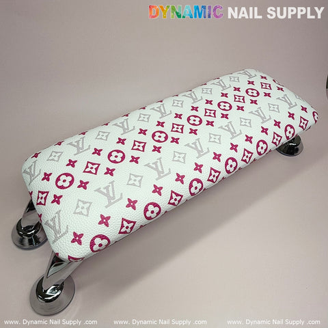 A rectangular footrest with a white and pink LV pattern design rests on four chrome legs. Styled like a leather pillow cushion, the upholstery showcases stylized letters and geometric symbols. The product, named the "(White Pink LV Pattern) ArmRest with Leather Pillow Cushion for Manicure Nails Service," is perfect for manicure enthusiasts and is offered by Dynamic Nail Supply.