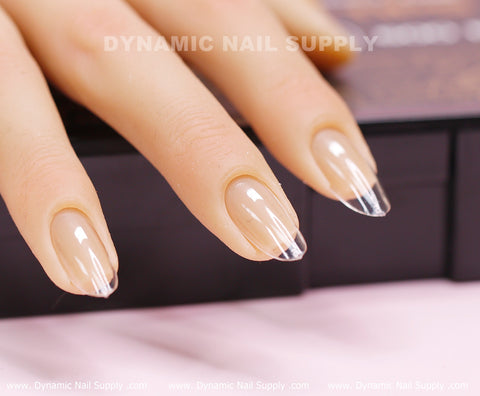 A close-up showcases a hand adorned with long, clear acrylic nails featuring Dynamic Nail Supply's [Round M] Full Cover Tips - Soft Gel X tips. The sophisticated nails possess a glossy finish and are elegantly applied over the natural nail bed, highlighting their transparent tips. The hand gently rests on a dark surface, complemented by the background text reading Dynamic Nail Supply.