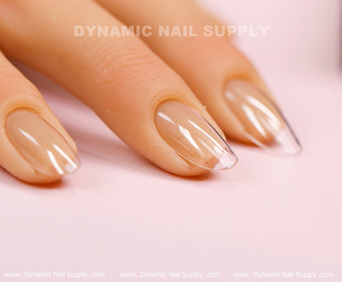 A close-up of a hand displaying neatly manicured nails with transparent soft gel tips, set against a light pink background. The medium-long, full cover tips from the [Round M] Soft Gel X collection showcase an almond shape with a glossy finish. The words Dynamic Nail Supply are faintly visible above the nails, repeated across the image.