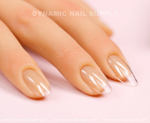 Manicured hands showcase clear, medium-long soft gel tips shaped to a point, accentuating the design from Dynamic Nail Supply's [Round M] Full Cover Tips collection. The nails exhibit a subtle nude shade at the base that elegantly transitions to a transparent tip as the fingers rest on a light pink surface. In the background, faint text reads: Dynamic Nail Supply.