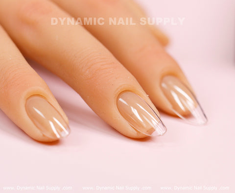 A close-up captures a hand adorned with medium long, clear acrylic nails featuring a subtle nude gradient using [Round M] Full Cover Tips - Soft Gel X tips by Dynamic Nail Supply. The glossy, pointed nails stand out against a soft pink backdrop, with the brand name prominently displayed above. This image highlights a professional manicure with a contemporary design.