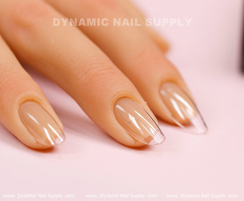A close-up showcases a hand with long, almond-shaped nails adorned with a clear base and beige tips. The glossy, well-manicured appearance is achieved using Dynamic Nail Supply's [Round M] Full Cover Tips - Soft Gel X tips. Positioned against a soft pink backdrop, the brand name "Dynamic Nail Supply" subtly appears at the top of the image.