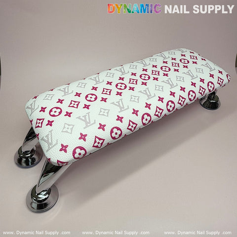 A white leather pillow cushion armrest for manicures, with silver legs and a designer-inspired pink pattern on a soft beige background. The space-saving armrest is topped with "Dynamic Nail Supply" in colorful letters, while the bottom displays a repeating company watermark.