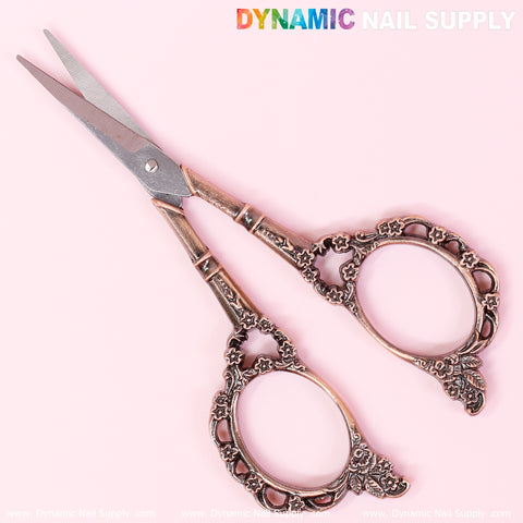 A set of multi-functional stainless steel nail scissors by Dynamic Nail Supply, designed for manicure tasks, is showcased against a soft pink background. These scissors feature ornate handles with circular loops and intricate floral patterns, enhancing their elegant and vintage-style aesthetic.