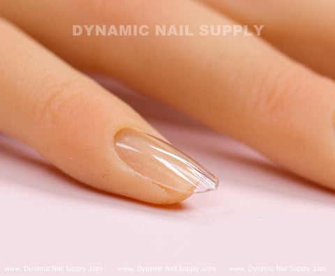 A close-up of a mannequin hand showcasing Round M Full Cover Tips by Dynamic Nail Supply, featuring clear, glossy acrylic nail extensions set against a soft pink background. The medium long, elegantly shaped nails resemble stylish press-on nails with a smooth, shiny finish. The background displays the repeating text "Dynamic Nail Supply" in light gray.