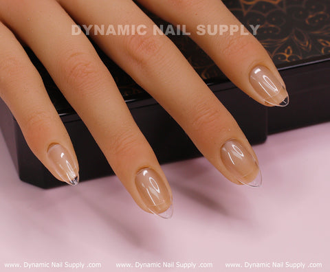 A mannequin hand with long, oval Soft Gel X tips is positioned on a black rectangular base. The clear, glossy nails are meticulously applied, showcasing the natural hue of the mannequin's fingertips. Behind it lies a patterned dark brown surface. At the bottom is branding text that reads Dynamic Nail Supply.