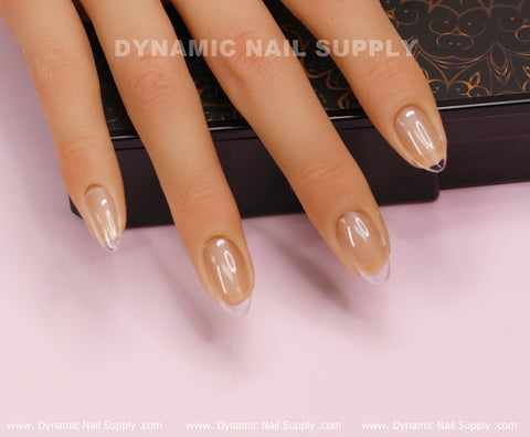 A mannequin hand elegantly displays semi-transparent nude almond-shaped nails adorned with [Oval] Full Cover Tips made from soft gel. These regular length tips are available in 11 sizes and come in a set of 600 pieces. The nails rest atop a black box featuring floral patterns, all set against a light pink background. The name "Dynamic Nail Supply" is prominently printed across the top and bottom of the scene.