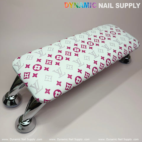 This compact armrest, the (White Pink LV Pattern) ArmRest with Leather Pillow Cushion for Manicure Nails Service by Dynamic Nail Supply, features a white padded resting surface adorned with luxurious pink and gray patterns. It is supported by four elegant silver metallic legs. The branding text, "Dynamic Nail Supply," is displayed in a gradient and white font against a light cream background.