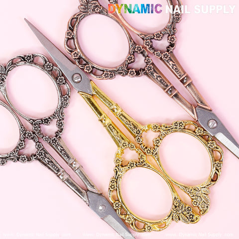 On a pink background, three ornate pairs of embroidery scissors are displayed. Two pairs feature silver stainless steel with floral designs, while one pair showcases a gold finish with equally intricate patterns. These scissors boast an elegant vintage aesthetic with looped handles. The brand name, "Dynamic Nail Supply," is prominently visible at the top.