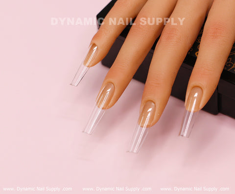 A realistic mannequin hand rests on a pink surface, showcasing the [Square L] Full Cover Tips from Dynamic Nail Supply. These clear acrylic nails are neatly shaped in a long length and extend beyond the fingertips. The image features text that reads Dynamic Nail Supply at the top.