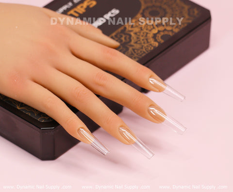 A realistic mannequin hand featuring long, clear acrylic nails in a Square Long shape is displayed on a black and gold patterned box. The box, branded with Dynamic Nail Supply, is set against a soft pink backdrop, elegantly highlighting the allure of the [Square L] Full Cover Tips - Soft Gel X tips.