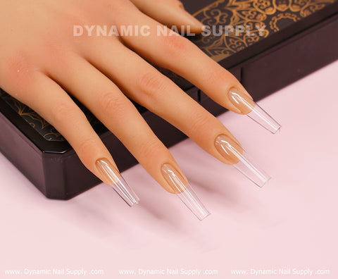 A realistic mannequin hand rests on a black and gold patterned box, showcasing full cover, clear soft gel X tips from Dynamic Nail Supply. The long-length Square L tips are prominently displayed on the palm-down hand, emphasizing their length and clarity. The scene is enhanced by a soft pink background with the Dynamic Nail Supply text visible.