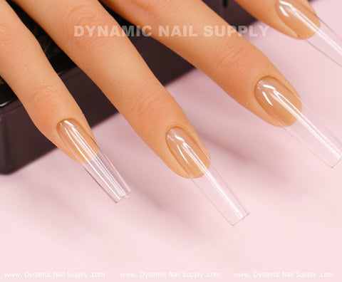 An image featuring a close-up of a mannequin hand showcases long, clear, square-shaped nails from the [Square L] Full Cover Tips - Soft Gel X tips collection by Dynamic Nail Supply. Set against a soft pink background, the company's watermark, DYNAMIC NAIL SUPPLY, is displayed across the picture. The full cover tips exhibit a glossy finish and precise shaping.