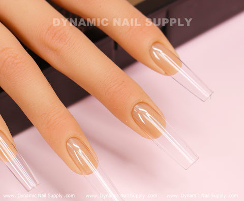 A close-up features a hand displaying long, rectangular clear nails with a nude base, shaped in the distinctive square long style. These elegant nails are crafted using the [Square L] Full Cover Tips from Dynamic Nail Supply. The brand name, Dynamic Nail Supply, appears subtly in the background as the hand is poised over a black surface against a gentle pink backdrop.