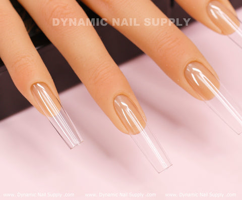 A close-up shot features a hand with long, square-shaped acrylic nails against a light pink background. The glossy, clear nails reveal the natural ones beneath. At the top, the text reads Dynamic Nail Supply, highlighting their [Square L] Full Cover Tips - Soft Gel X tips for a perfect blend of style and elegance.