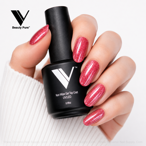 A hand with sparkly red nails holds a V Beauty Pure - Non Wipe Gel Top Coat bottle against a white background.