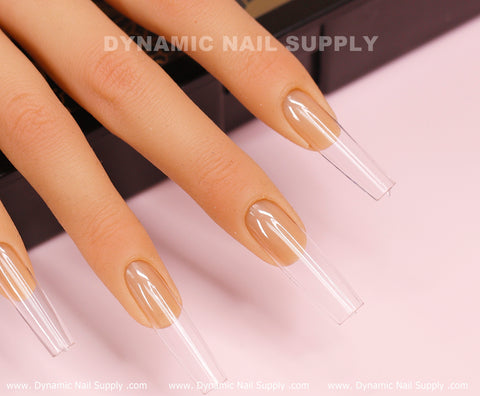 A close-up of a hand with three fingers displays long, clear extensions using the "Square L" Full Cover Tips—Soft Gel X tips by Dynamic Nail Supply. Neatly applied with a natural finish in a square long shape, they stand out against a soft pink background. The image includes "DYNAMIC NAIL SUPPLY" at the top and website details at the bottom.