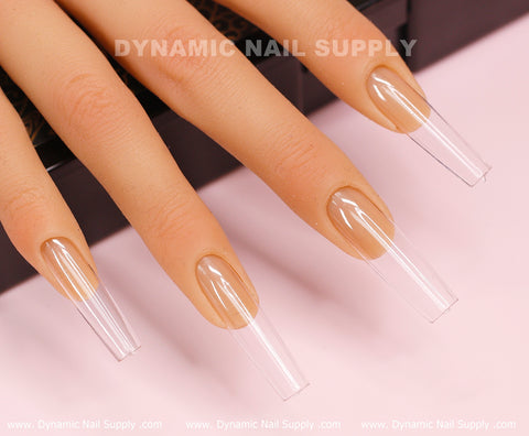 The image showcases a close-up of a mannequin hand adorned with long, transparent acrylic nails styled in a sleek square shape. The light pink background enhances the elegance of the [Square L] Full Cover Tips - Soft Gel X tips from Dynamic Nail Supply, which are neatly shaped and glossy to highlight their modern appeal. The text "DYNAMIC NAIL SUPPLY" appears at both the top and bottom of the image.