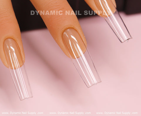 A close-up of a mannequin hand showcases three semi-transparent [Square L] Full Cover Tips - Soft Gel X tips in a long length, extending beyond the natural nails. The clear full cover tips feature a contemporary squared design against a light pink background that highlights the nail art. The branding text "Dynamic Nail Supply" is visible at the top edge.