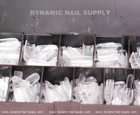 Clear plastic nail tips, specifically the Square M Full Cover Tips - Soft Gel X tips in medium length, are neatly housed in a gray storage container with multiple compartments. Each compartment stores different sizes from the set of 11. "Dynamic Nail Supply" is prominently featured at the top, while text including their website details decorates the bottom.