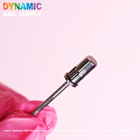 A close-up of a person wearing a pink glove holding an Easy-Off Sanding Band Mandrel against a pink background. The high-quality metal bit features grooves, designed for use with any sanding band. The Dynamic Nail Supply logo is visible in the top left corner, showcasing the tool's details.