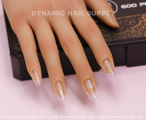 A mannequin hand presents a set of [Square M] Full Cover Tips - Soft Gel X tips, medium length and clear with square tips, on an ornately patterned box. The backdrop is a light pink surface, featuring the Dynamic Nail Supply brand prominently on both the box and surface.