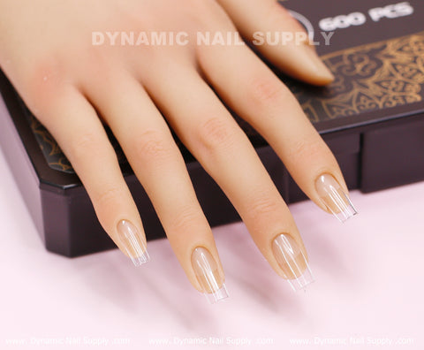 A hand model showcases impeccably manicured fingers adorned with Soft Gel X tips from the [Square M] Full Cover Tips collection, featuring clear, square-tipped nails. Resting on a decorative box, the text "DYNAMIC NAIL SUPPLY" is prominently displayed in white against a soft pink background.