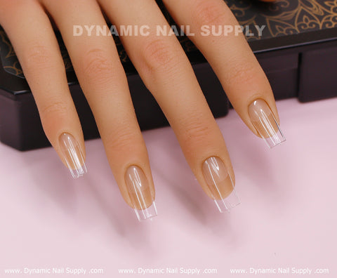 Close-up of a realistic hand model showcasing Medium Length Square M Full Cover Tips made from soft gel, highlighting the elegant transition from glossy beige to transparent tips. The scene is set against a decorative black box backdrop, prominently displaying "Dynamic Nail Supply" both in the foreground and background.