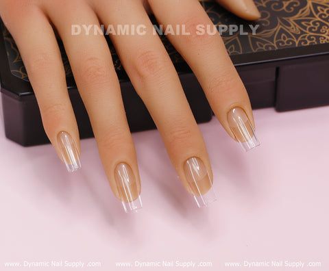 A mannequin hand presents the [Square M] Full Cover Tips from Dynamic Nail Supply, with long, transparent Medium Square Nail Tips set on a decorative black and gold-patterned box. The tips exhibit a smooth, glossy finish. Text displaying "DYNAMIC NAIL SUPPLY" is placed along the top and bottom borders of the image.