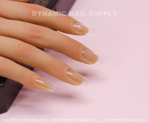 A realistic mannequin hand is displayed with [Square M] Full Cover Tips from Dynamic Nail Supply. These Medium Length Soft Gel X tips feature a transparent, glossy finish and are neatly filed to extend just beyond the fingertips. The hand is placed on a light pink surface, with "Dynamic Nail Supply" printed in white in the background.