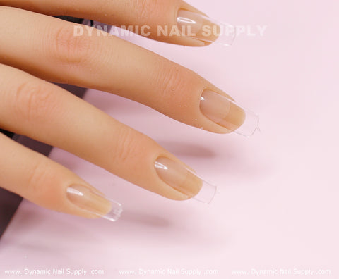 Close-up of manicured fingers displaying transparent acrylic nail extensions on a light pink surface. The nails, crafted with [Square M] Full Cover Tips in Medium Length, show off a glossy finish. "Dynamic Nail Supply" is prominently displayed at the top of the image, and their website is conveniently located at the bottom for easy access.