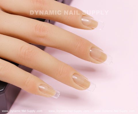 A close-up of a realistic hand model highlights the glossy, transparent [Square M] Full Cover Tips - Soft Gel X tips by Dynamic Nail Supply. The neatly manicured nails extend slightly beyond the fingertips, boasting a shiny finish. The background subtlety displays the text 'Dynamic Nail Supply' while the hand rests gracefully on a dark surface.