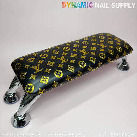 A luxurious manicure armrest featuring a leather cushion with a black and gold LV pattern, complemented by four shiny silver legs. The Dynamic Nail Supply brand is prominently displayed at the top. Perfect for any manicure service, its design stands out against a neutral background.