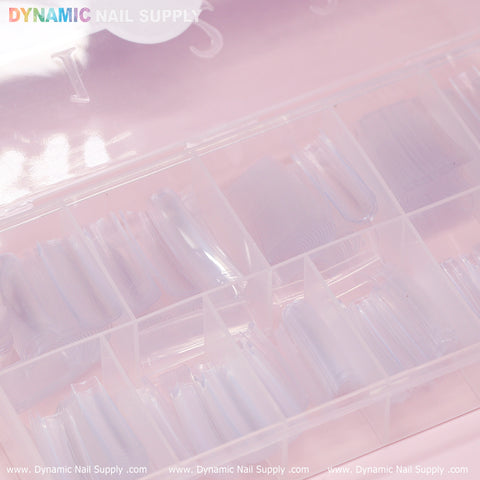 A clear plastic box with organized compartments in a pastel pink setting showcases durable, transparent artificial nail tips. The lid features the words "Dynamic Nail Supply" in vibrant lettering, along with the brand's website at the bottom edge. Ideal for XXL Tapered Square Tips (New 2024) - XXL - 11 sizes - 550 tips - Clear.