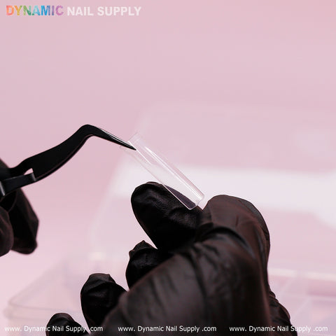 A person wearing black gloves holds a durable, clear XXL Tapered Square Tip with black tweezers. The light pink background features the colorful text "Dynamic Nail Supply" at the top left, while the repeated text "www.DynamicNailSupply.com" appears at the bottom.