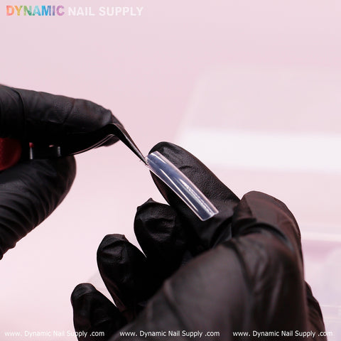 A person wearing black gloves delicately holds a clear acrylic nail extension with tweezers, showcasing the durable quality of XXL Tapered Square Tips (New 2024). The soft pink background highlights Dynamic Nail Supply at the top, with www.DynamicNailSupply.com elegantly repeated along the bottom edge.