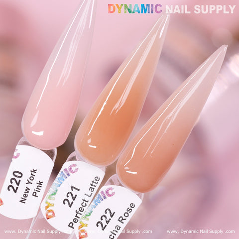 A close-up of three acrylic nail swatches from Dynamic Nail Supply showcases the Nude Acrylic Powder for durable nails. From left to right, the shades are New York Pink (#220) in light pink, Perfect Latte in medium beige, and Vienna Rose in soft peach. The long, pointed swatches are set against a softly blurred pink background.