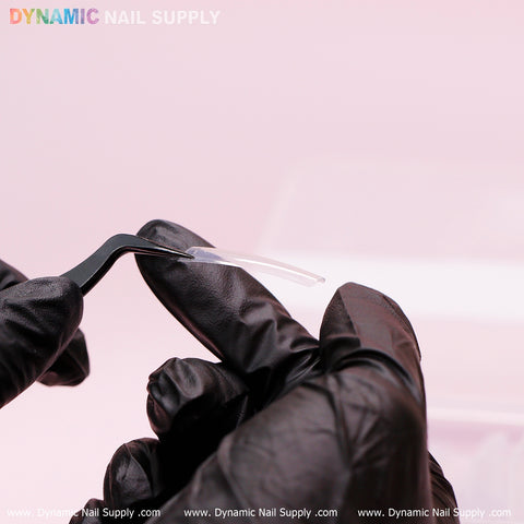 A gloved hand displays a black nail tool and an XXL Tapered Square Tip against a pale pink background. The image highlights Dynamic Nail Supply's branding at the top left and along the bottom edge in colorful text, showcasing their durable XXL Tapered Square Tips (New 2024) in clear, designed for professionals.