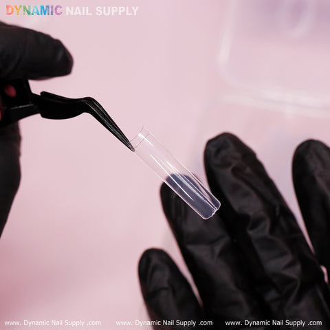 A person wearing black gloves uses black tweezers to hold an XXL Tapered Square Tip against a soft pink background. At the top, the words "Dynamic Nail Supply" appear in colorful text, showcasing their durable clear tips from the new 2024 collection.