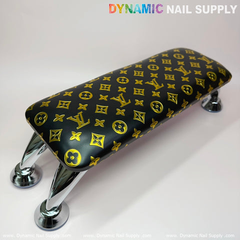 A black, leather cushion armrest with a pillow, featuring a gold LV pattern reminiscent of a luxury brand logo, is supported by four shiny, curved chrome legs, ready for manicure service. The background is plain white. At the top, the text Dynamic Nail Supply is showcased in colorful lettering.