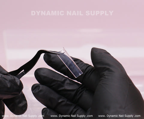 A gloved hand uses tweezers to apply XL Square Nail Tips (Not C-Curve) on another hand against a pink background.