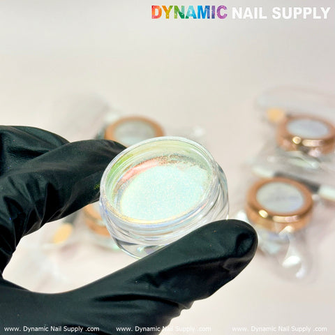 A gloved hand holds a small, open container of Golden/Light-Orange Iridescent Chrome #02 - High brightness Fine Pigment Powder, highlighting its shimmering surface. In the background, several similar closed containers with rose gold lids are slightly blurred. The text Dynamic Nail Supply appears at the top of the image, suggesting endless possibilities for perfect nail art designs.
