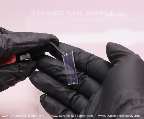 A gloved hand uses tweezers to apply XL Square Nail Tips – Extra Long Straight Shape, with branding in the background.