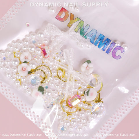The Luxury White Pearls and Flowers with Leaves Set (White Theme) for Nail Art Design from Dynamic Nail Supply is presented on a pink background. This collection features luxury white pearls, gold-colored crescent moons, and rose-shaped embellishments, all neatly arranged in a sealed package adorned with a decorative white bow. The top of the package showcases the word "DYNAMIC" in rainbow letters.